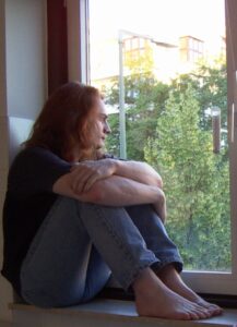 A nerd with long shaggy red hair perched in a deep window frame in an apartment, looking pensively out at the sunny world outside.  It was actually really uncomfortable sitting like that and I only did it because I thought it would make a cute selfie.